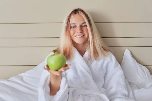 Жінка-підліток з зеленим яблуком у білому халаті сидить у ліжку — стокове фото