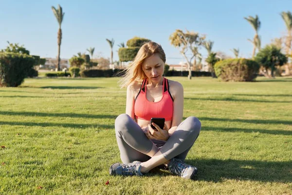 성숙 한 여성 이 녹색 잔디에 대한 아침 운동을 하고 있습니다. 스마트폰 과 비디오 운동을 하고 있죠. — 스톡 사진