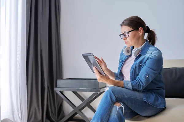 Seriöse Frau mittleren Alters im Kopfhörer mit digitalem Tablet zu Hause auf der Couch — Stockfoto