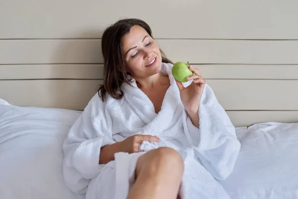 Middelbare vrouw in witte badjas met groene appel in bed — Stockfoto