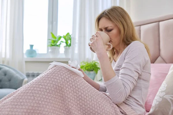 Mujer de mediana edad enferma sentada en casa en la cama con pañuelo y taza — Foto de Stock