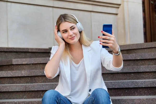 Mladá krásná žena ve sluchátkách s chytrým telefonem odpočívá na schodech ve městě — Stock fotografie