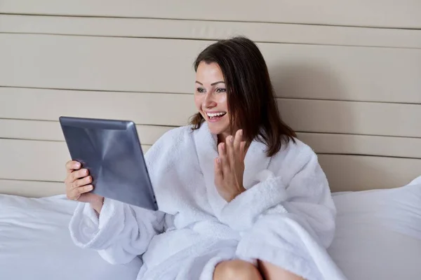 Mulher madura em roupão de banho branco olhando para webcam de tablet digital sentado na cama, falando online — Fotografia de Stock