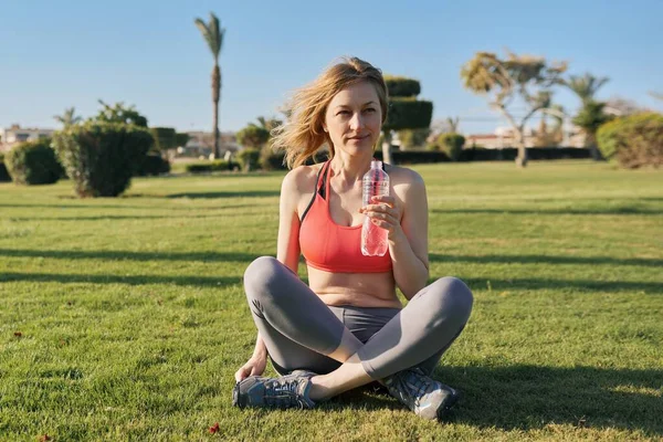 Ώριμη γυναίκα κάνει πρωινές ασκήσεις σε πράσινο γρασίδι στο πάρκο, με ένα μπουκάλι νερό — Φωτογραφία Αρχείου
