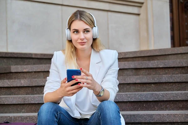 Giovane bella donna in cuffia con smartphone seduto su gradini in città — Foto Stock