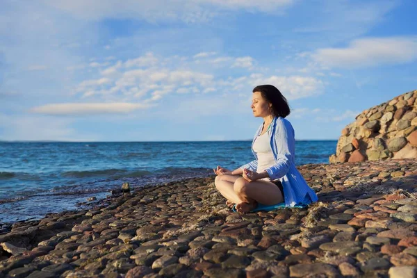 Красивая женщина средних лет, сидящая в позе лотоса медитируя на берегу моря на закате — стоковое фото