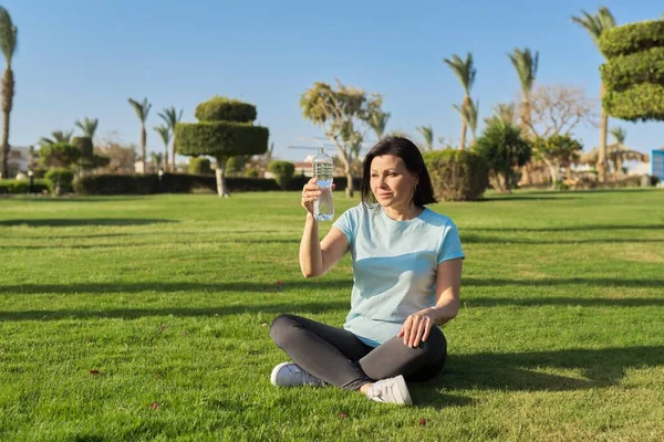 Mujer madura haciendo ejercicios matutinos sobre hierba verde en el parque, con botella de agua — Foto de Stock