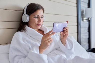 Bornoz ve kulaklıklı bir kadın akıllı telefon ekranına bakıyor, yatakta oturuyor.