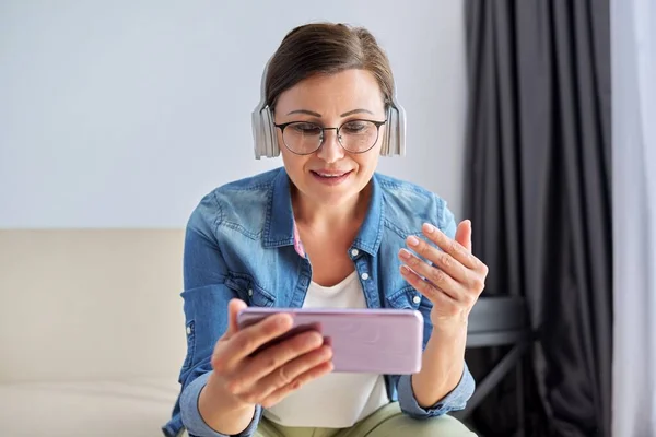 Mujer de mediana edad con auriculares y mirando la pantalla del teléfono inteligente sonriendo hablando —  Fotos de Stock