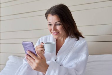 Bornozlu, elinde bir fincan kahveyle gülümseyen bir kadın akıllı telefon ekranına bakıyor, yatakta oturuyor.