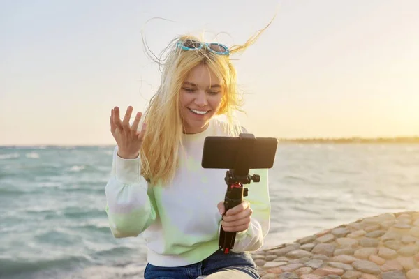 Female teenager with smartphone on tripod on sea sunset beach recording video