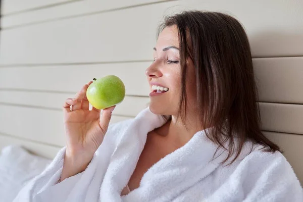 Жінка середнього віку в білому халаті з зеленим яблуком у ліжку — стокове фото