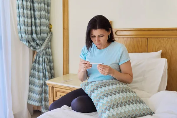 Sorglig sjuk mogen kvinna som håller termometer i sina händer, sitter hemma i sängen — Stockfoto