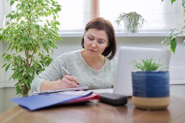 Evde dizüstü bilgisayar kullanarak çalışan olgun bir kadın.