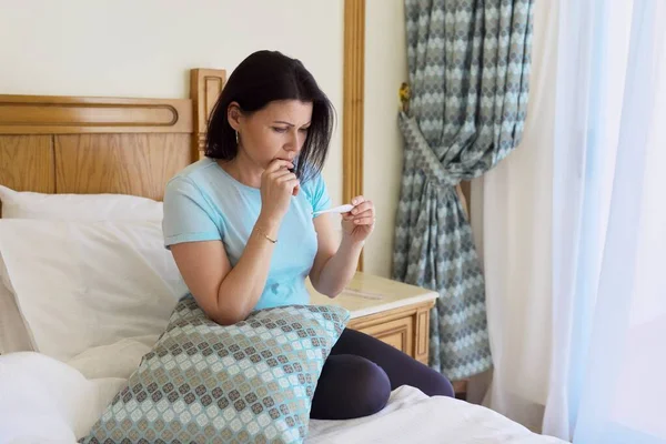 Traurig kranke reife Frau hält Thermometer in der Hand, sitzt zu Hause im Bett — Stockfoto