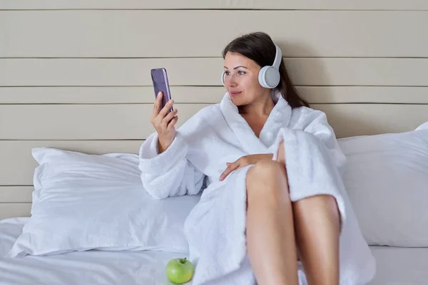 Mulher madura feliz em roupão de banho branco e fones de ouvido com smartphone sentado na cama — Fotografia de Stock