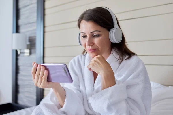 Mujer en albornoz y auriculares mirando la pantalla del teléfono inteligente, sentado en la cama —  Fotos de Stock