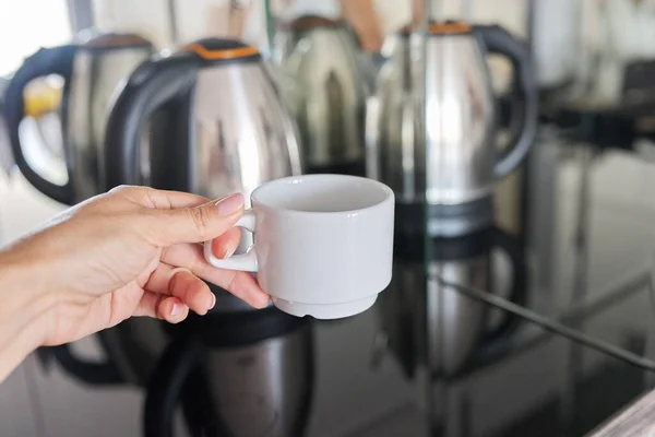 Bílý šálek kávy v ruce close-up, nerezová ocel rychlovarná konvice v zrcadle pozadí — Stock fotografie