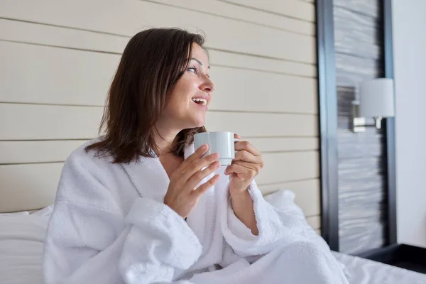 Mooie volwassen vrouw met kopje koffie in bed in witte badjas zitten in bed — Stockfoto