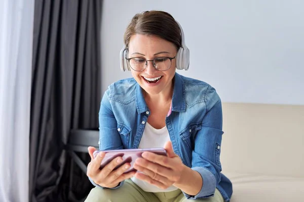 Middle aged woman with headphones and looking at smartphone screen smiling talking — Stock Photo, Image