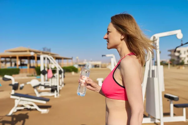 Mujer madura haciendo ejercicio deportivo en simuladores al aire libre, con botella de agua — Foto de Stock