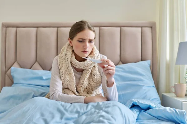 Άρρωστη νεαρή γυναίκα με θερμόμετρο στα χέρια της, κάθεται στο σπίτι στο κρεβάτι — Φωτογραφία Αρχείου
