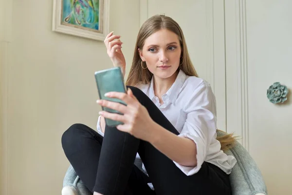 年轻的金发姑娘坐在家里的椅子上，用智能手机进行视频交流 — 图库照片