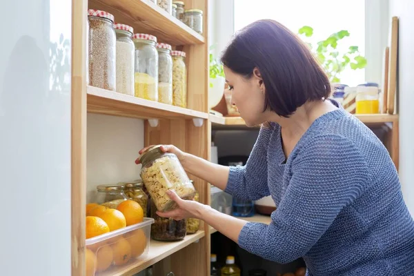 Organization of pantry, woman in kitchen near wooden rack with cans and containers of food