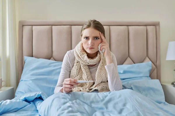 Mujer joven enferma con termómetro en las manos, sentada en casa en la cama —  Fotos de Stock