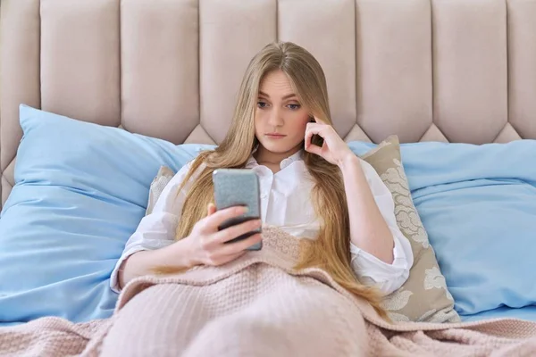 진지하게 놀란 젊은 여성 이 스마트폰을 들고 침대에 누워 책을 읽고 집중하는 모습 — 스톡 사진