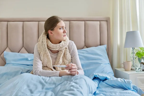 Mujer joven enferma con termómetro en las manos, sentada en casa en la cama — Foto de Stock