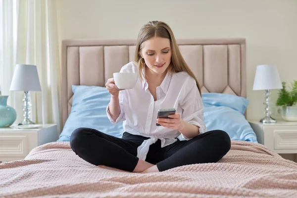 Mladý usmívající se blondýnka s šálkem kávy a smartphone sedí v posteli — Stock fotografie