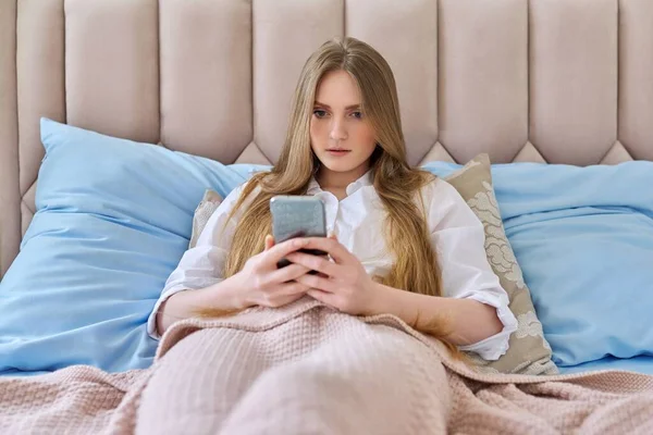 스마트폰을 들고 집에서 침대에 누워 쉬고 있는 진지 한 젊은 여성, — 스톡 사진