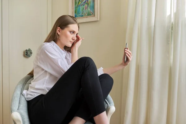 집에서 의자에 앉아 있는 금발의 어린 소녀, 스마트폰으로 비디오 통신을 사용하는 모습 — 스톡 사진