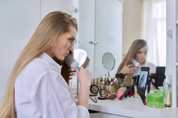 Jovem mulher sentada na mesa de maquiagem com espelho fazendo maquiagem e penteado — Fotografia de Stock