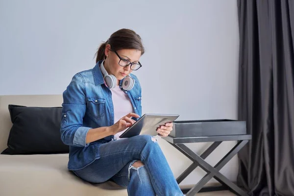 Mujer seria de mediana edad en auriculares con tableta digital en casa en el sofá —  Fotos de Stock