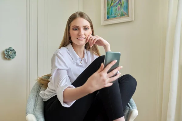 Mladá blondýna sedí doma v křesle, pomocí video komunikace na smartphonu — Stock fotografie