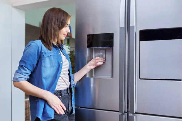Water dispenser, woman taking cold water into glass from home refrigerator