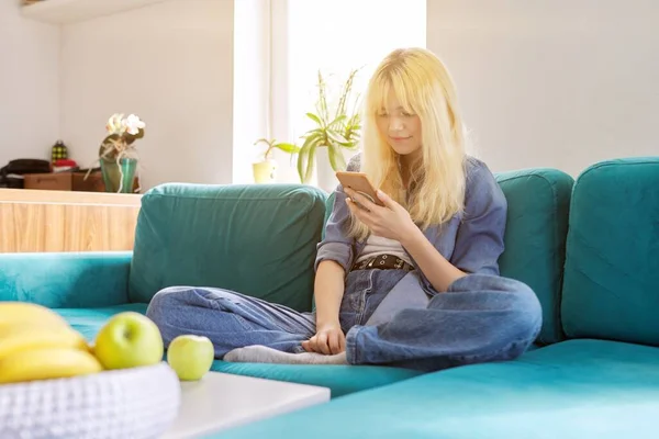 Усміхнена щаслива жінка-підліток сидить вдома на дивані, дивлячись на екран смартфона — стокове фото