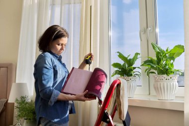 Female designer choosing curtain fabrics, measuring window with tape measure