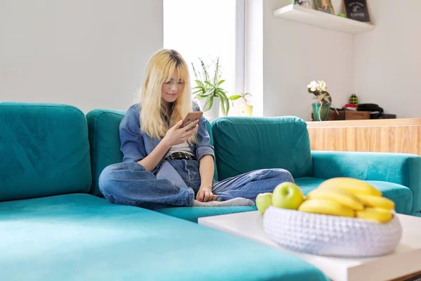 Улыбающаяся счастливая девушка-подросток сидит дома на диване и смотрит на экран смартфона — стоковое фото