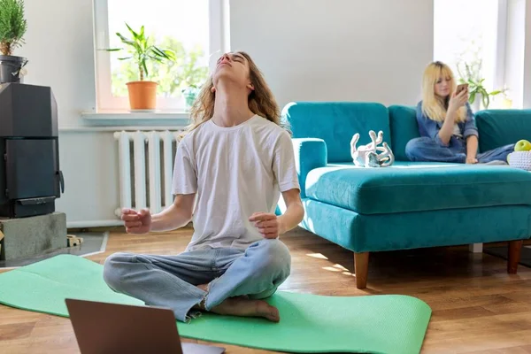 Guy teenager sedí v lotosové pozici na podložce jógy doma na podlaze s laptopem — Stock fotografie