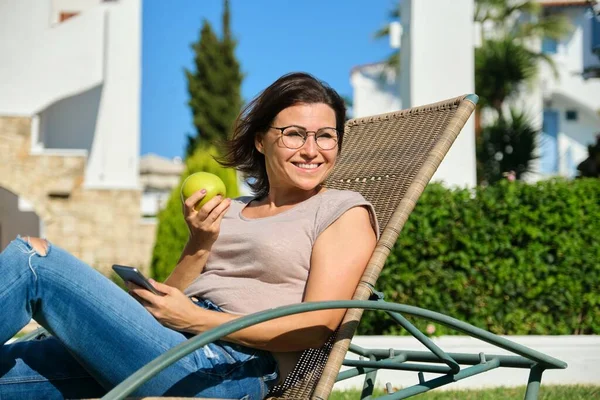 Šťastná fena sedí v zahradním křesle s jablkem a smartphonem na zeleném trávníku — Stock fotografie