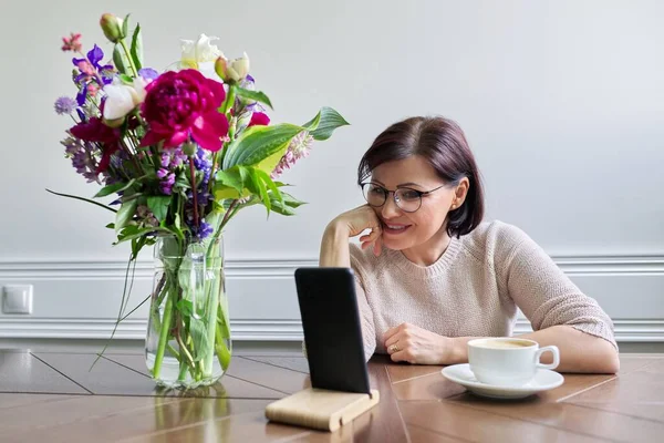 Mujer de mediana edad sentada en la mesa en casa mirando la pantalla del teléfono inteligente —  Fotos de Stock
