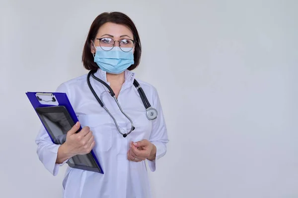 Retrato de doctora en mascarilla médica con portapapeles y tableta digital, sobre fondo claro — Foto de Stock