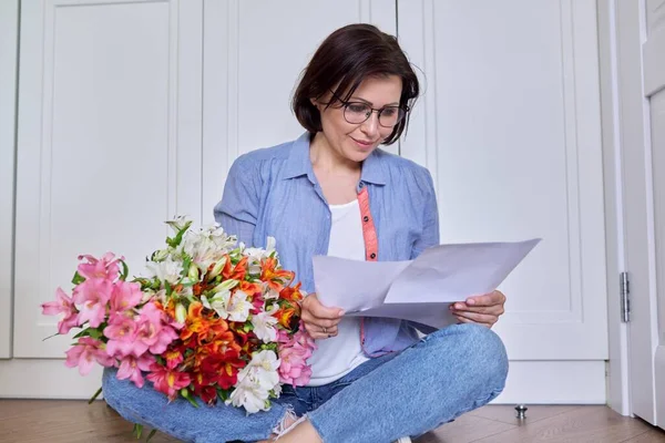 Улыбающаяся женщина средних лет с букетом цветов, читающая бумагу — стоковое фото
