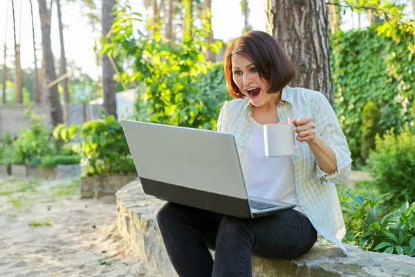 Beautiful emotional middle aged woman relaxing in garden with cup of tea and laptop