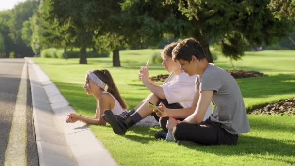 Hipster teenagers having fun in park, sitting on green grass on lawn, recording videos on smartphone — Stock Video