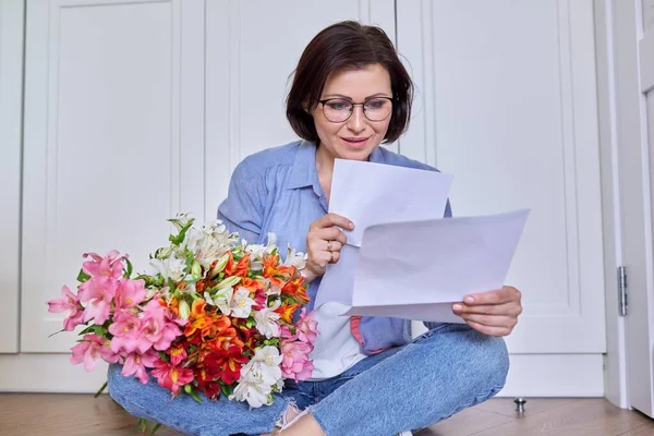 Улыбающаяся женщина средних лет с букетом цветов, читающая бумагу — стоковое фото