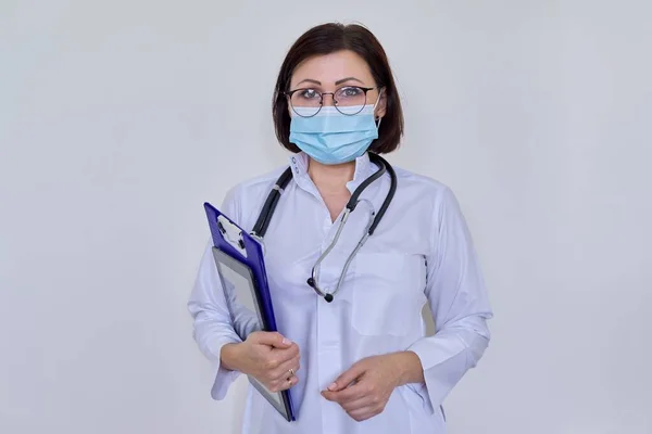 Retrato de doctora en mascarilla médica con portapapeles y tableta digital, sobre fondo claro — Foto de Stock